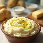 "Creamy mashed potatoes in a rustic bowl garnished with chives and melting butter, with ingredients in the background on a clean marble countertop"