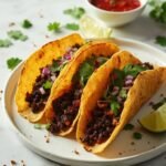 "Overhead view of crispy black bean tacos garnished with cilantro and onion on a clean white marble background, with fresh salsa and lime wedges in the background."