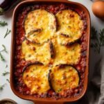 "Overhead view of a fresh eggplant and egg bake with melted cheese in a ceramic dish, surrounded by ingredients like whole eggplants, eggs, cheese, and oregano, against a neutral background."