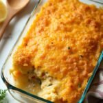 "Overhead shot of golden brown chicken casserole with crispy cracker topping and melted cheese in a baking dish on a clean white marble countertop, garnished with fresh parsley and a halved lemon"