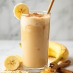 "Peanut butter banana smoothie garnished with banana wheel, cinnamon, and served with wooden straw, accompanied by fresh banana, peanut butter, rolled oats, and a cinnamon stick on a white marble surface with soft daylight"