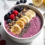 "Overhead view of a vibrant purple-pink smoothie bowl with banana slices, peanut butter, chia seeds, and granola on a clean white marble background, with frozen berries and half a banana slightly out of focus."