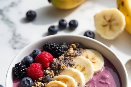 "Overhead view of a vibrant purple-pink smoothie bowl with banana slices, peanut butter, chia seeds, and granola on a clean white marble background, with frozen berries and half a banana slightly out of focus."