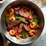"Overhead shot of steak and shrimp stir-fry with bell peppers and snap peas in a stainless steel wok, garnished with sesame seeds on a marble countertop."