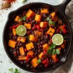 "Overhead view of a skillet with caramelized sweet potato cubes, black beans, and diced red bell peppers, garnished with cilantro and lime on a clean marble countertop"