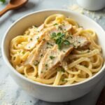 "Overhead shot of chicken alfredo pasta with Parmesan cream sauce in a white bowl on a marble countertop, with garnishes and extra cheese in the background."