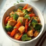 "Overhead view of a vibrant chicken and vegetable stir fry featuring broccoli, carrots, bell peppers in a white ceramic bowl with wooden chopsticks, on a clean white marble surface."