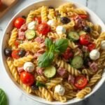 "Overhead view of classic pasta salad with rotini, cherry tomatoes, mozzarella, salami, and fresh basil in a white dish on a marble countertop"