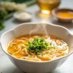 "Bowl of golden egg drop soup with silky ribbons, garnished with green onions and sesame oil on a clean marble countertop, with key ingredients in the background"