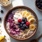 "Overhead view of hot oatmeal with mixed berries, banana slices and Greek yogurt in a rustic bowl on a clean marble surface, surrounded by ingredients like rolled oats and cinnamon."