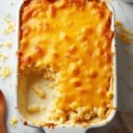 "Freshly baked homemade macaroni and cheese in a white ceramic dish on a marble background, with a serving spoon and extra grated cheese scattered around"