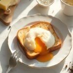 "Poached egg with runny yolk on buttered toast on a white plate, surrounded by cooking ingredients in warm morning sunlight"