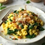 "Fluffy golden scrambled eggs mixed with wilted spinach and caramelized onions, topped with grated parmesan and red pepper flakes, served on a white ceramic plate against a clean white marble background."