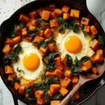 "Sweet potato hash with sunny-side-up eggs and wilted spinach in a cast iron skillet, served on a white marble background"