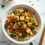 "Overhead view of vegetable and tofu stir-fry with brown rice in a white bowl on a marble surface, garnished with fresh parsley and accompanied by bamboo chopsticks and a ramekin of lemon wedges"